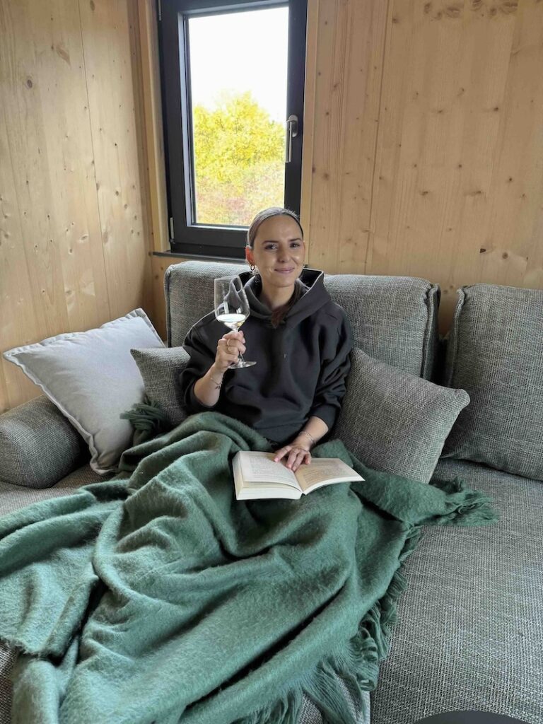 Sandra sitzt auf der Couch im Bocksbeutel Ferienhaus, liest ein Buch und trinkt Wein